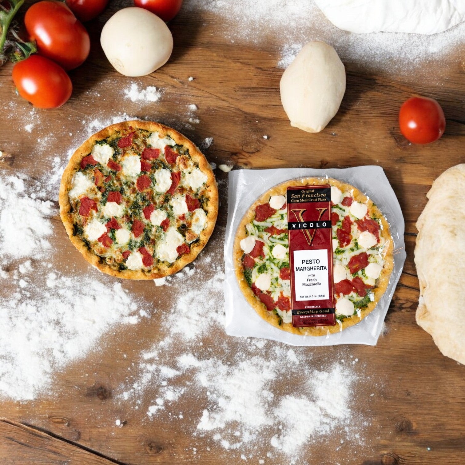 A packaged and an unwrapped Vicolo Pesto Margherita with Fresh Mozzarella pizza sit on a wooden surface, accompanied by tomatoes, dough balls, and flour.