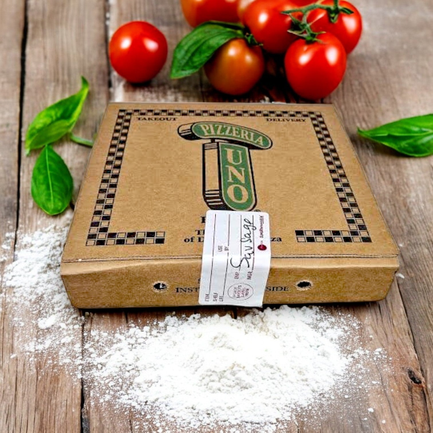 A Pizzeria Uno Deep Dish Small Sausage Pizza box on wood, amid fresh tomatoes, basil, and flour, evokes the delicious crafting of its renowned origins at Pizzeria Uno.