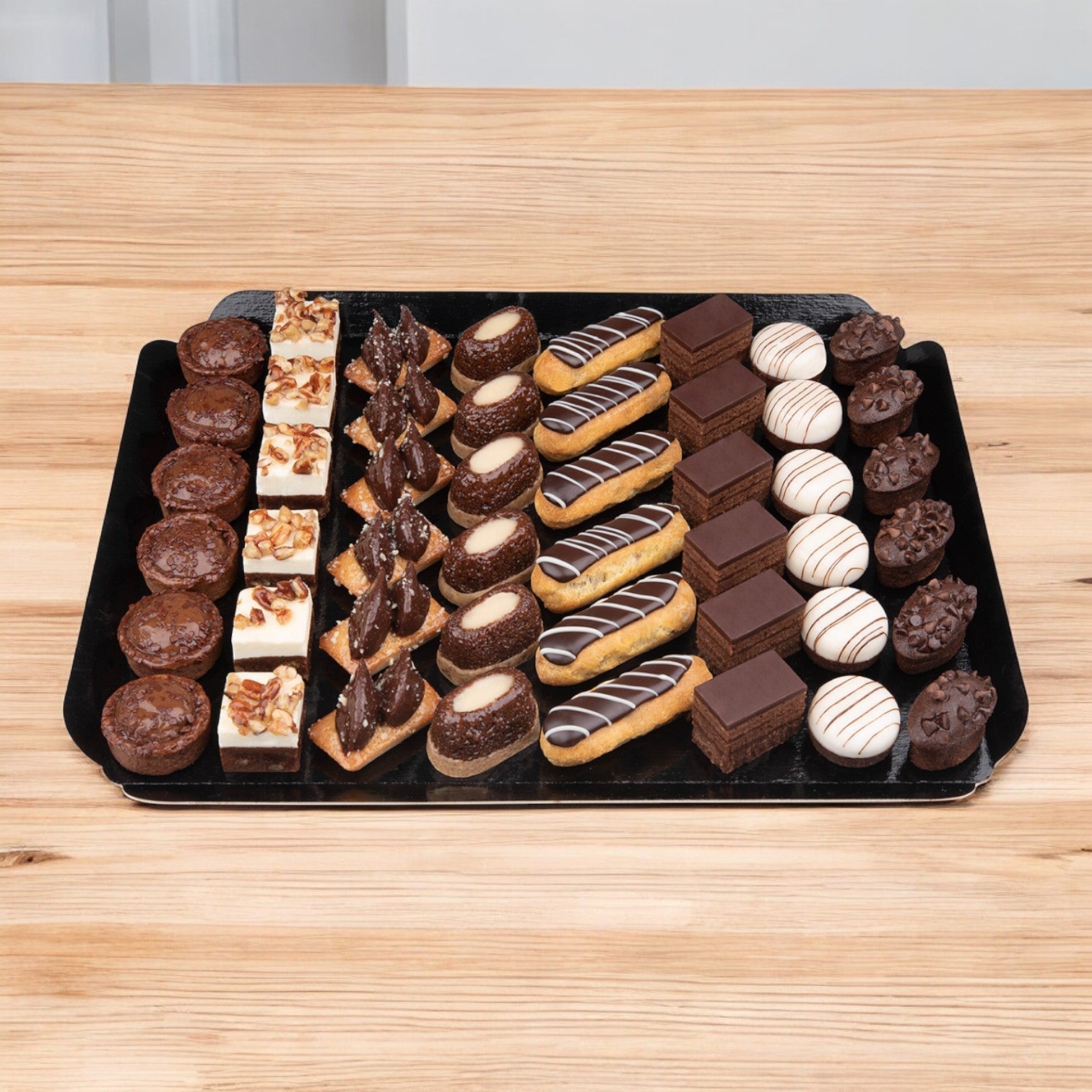 Pasquier's Cake Assortment Petits Fours Black & White 48 Piece offers a delightful display of French pastries, featuring eclairs, brownies, square-shaped cakes, and cream-filled treats elegantly presented on a black tray against a wooden surface.