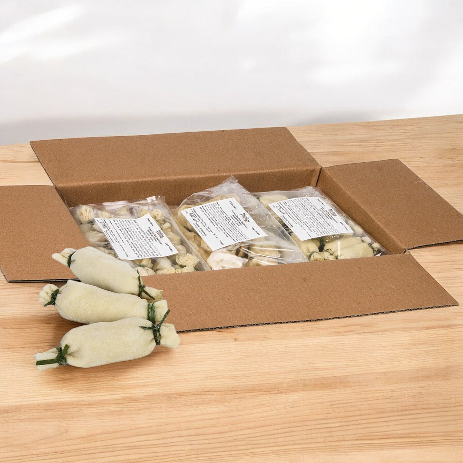A box of Phillips Foods Appetizer Dim Sum Fried sits open on a wooden table, revealing 72 pieces of delightful dim sum. Three bags inside boast authentic flavors, while four extra pieces are playfully scattered outside the box, tempting every gourmet.