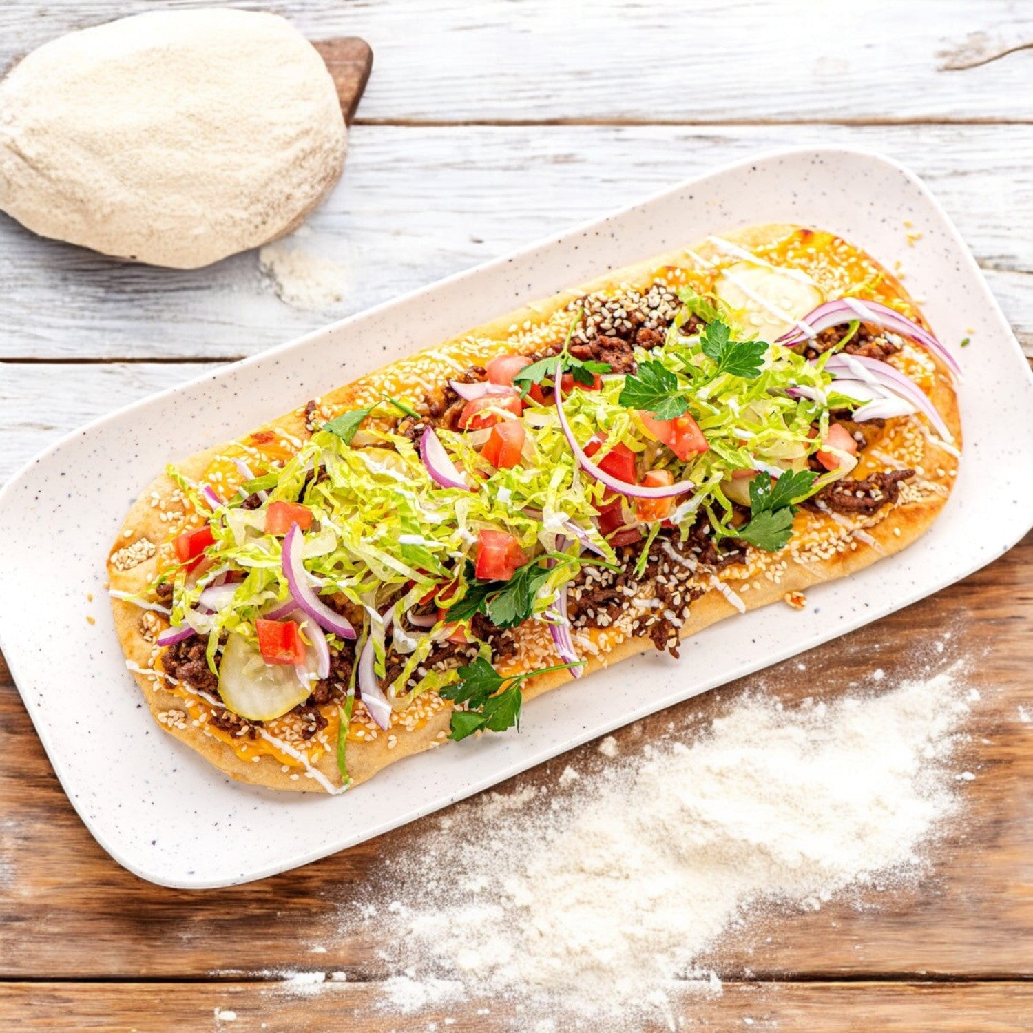 Rich’s Bread Flat Oval 12" X 5", from Rich's, adorned with crisp lettuce, juicy tomatoes, onions, and seeds rests on a white plate amidst scattered flour on the wooden table.