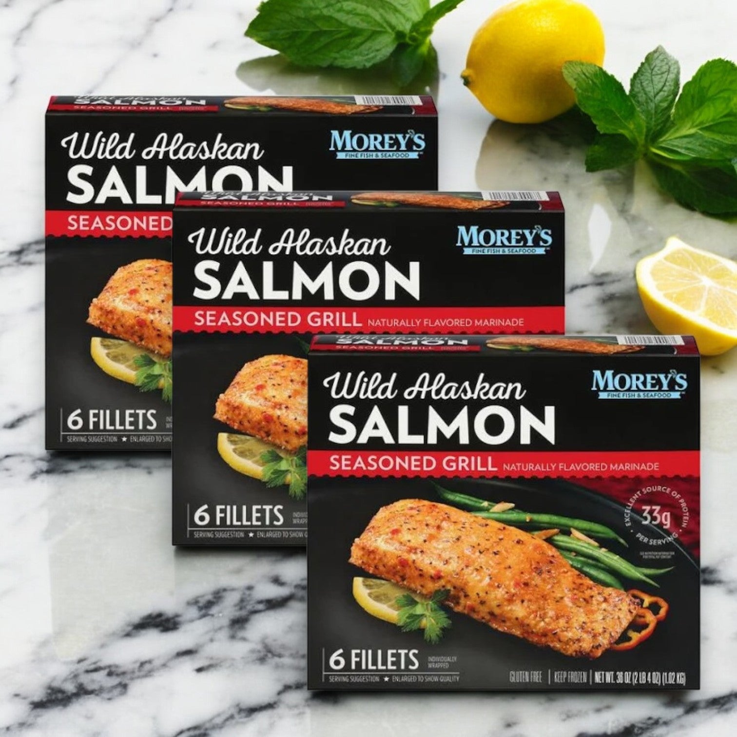 Three boxes of Morey's Marinated Wild Alaskan Salmon, 18 ct, from Morey's Fine Fish & Seafood are displayed on a marble surface, surrounded by fresh lemon and mint leaves.