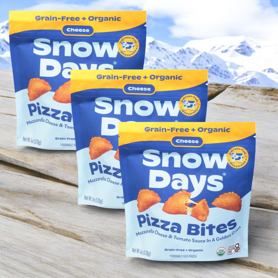 Three bags of Snow Days Frozen Organic Taco Pizza Bites are displayed on a wooden surface, with snow-covered mountains as a scenic backdrop.