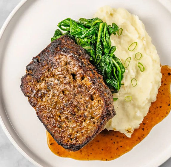 Experience the tender texture of FoodHaul Wagyu Beef Meatloaf, served with a side of mashed potatoes and wilted spinach. This premium frozen meal is topped with green onions and drizzled with gravy, presented elegantly on a white plate.